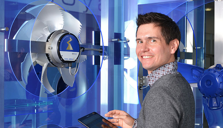 Jochen Brandt - Abteilungsleiter Entwicklung "Digitale Produkte" Junger lächelnder Mann steht mit einem Tablet vor einem ZIEHL-ABEGG Ventilator