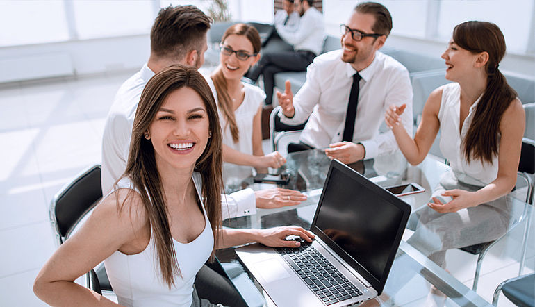 Personen bei der Arbeit am Laptop und bei einer Besprechung Lächelnde Person vor dem Laptop, im Hintergrund Personen bei einer Besprechung